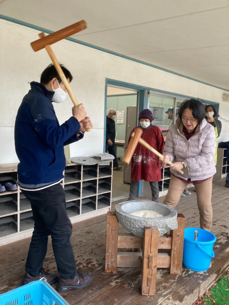 施設の縁側で、男女二人が臼を囲んで餅つきをしており、周囲で数人が見守っている