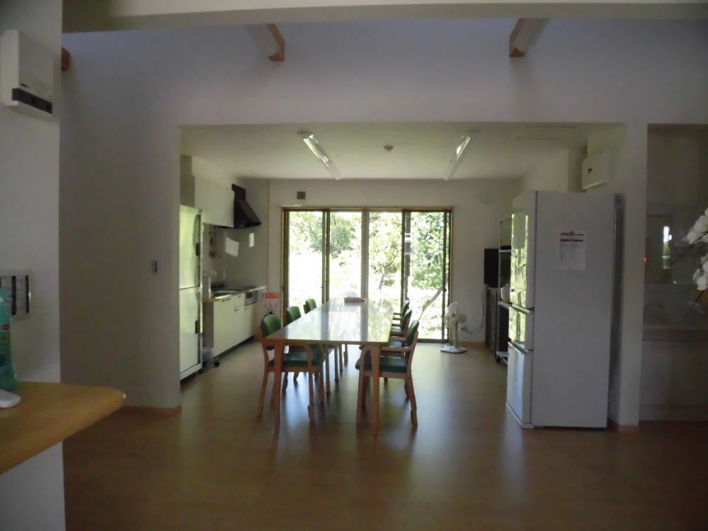 中央にテーブルと椅子6脚が置かれている。奥には広い掃き出し窓があり、木の緑が見える。左右にはキッチンや食器棚、冷蔵庫がある。