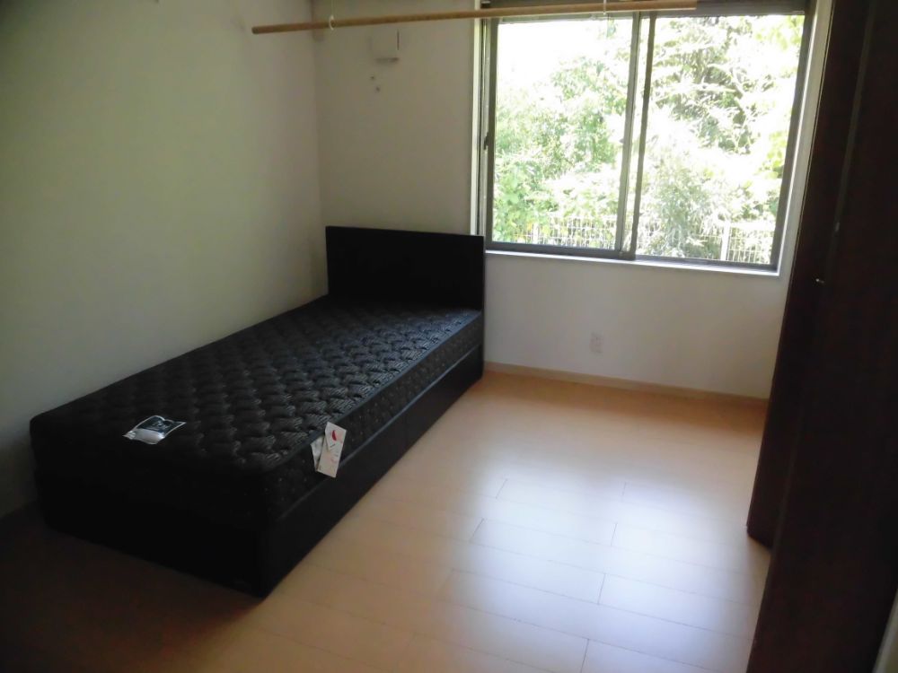 6畳ほどの板張りの居室。左手に黒いマットレス付きのベッドが置かれている。奥に窓があり、緑が見える