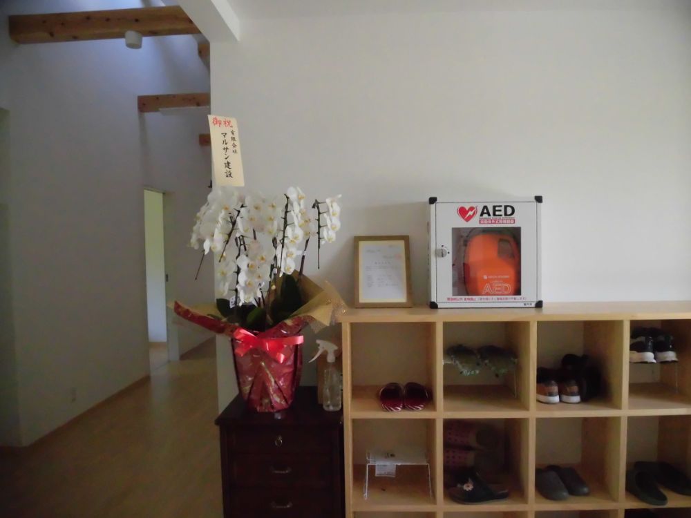 玄関内にある靴箱。写真では少なくとも12の収納スペースが見える。靴箱の上にAEDが置かれている。靴箱の横にはチェストがあり、その上には胡蝶蘭が飾られている